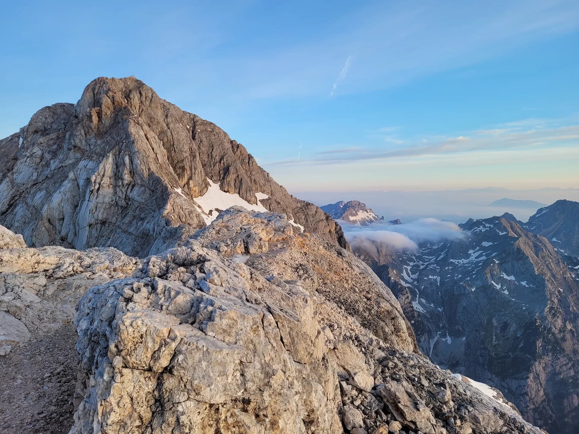 Rocky mountain ridge with snow patches, view towards Triglav summit and lower peaks above clouds.