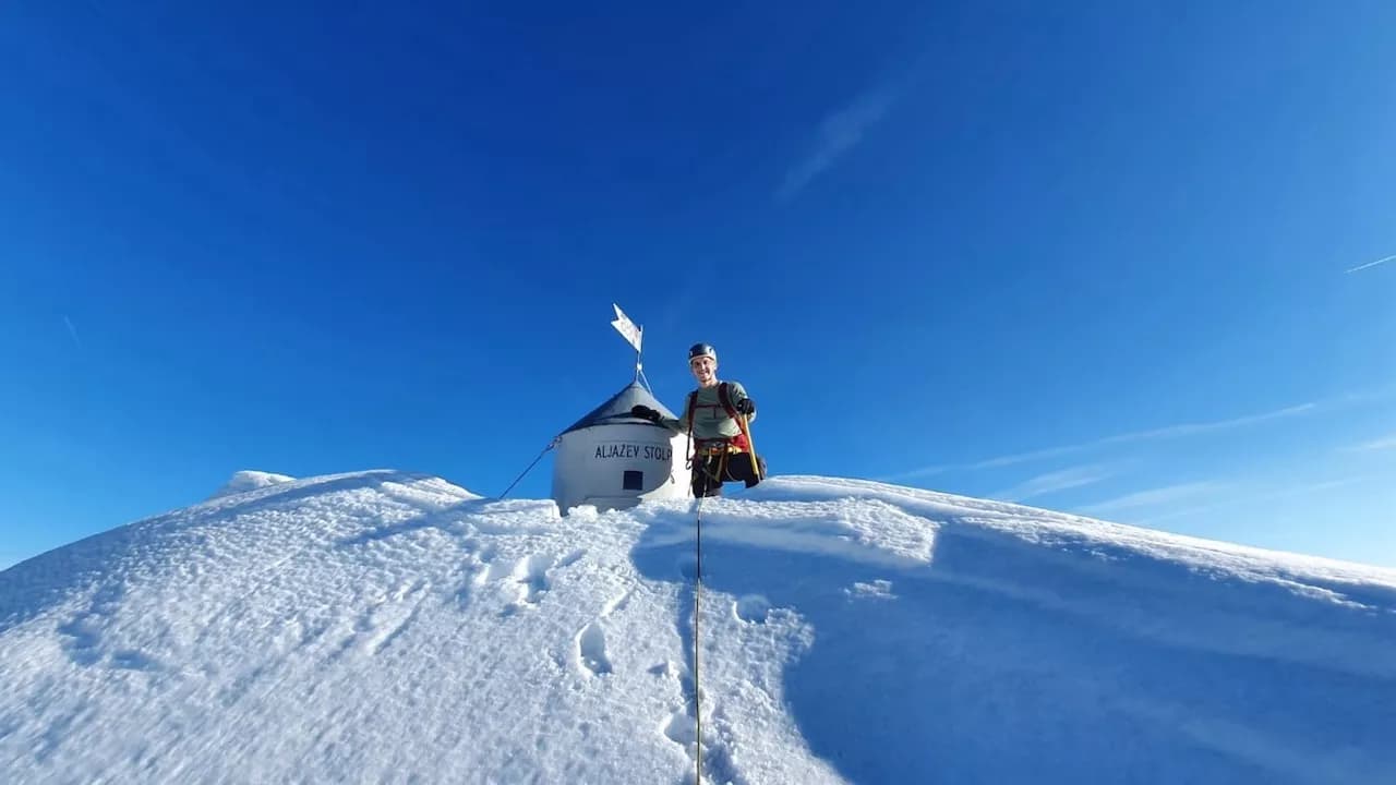 Mountaineer by Aljažev Stolp summit marker on deep snow under bright blue winter sky.