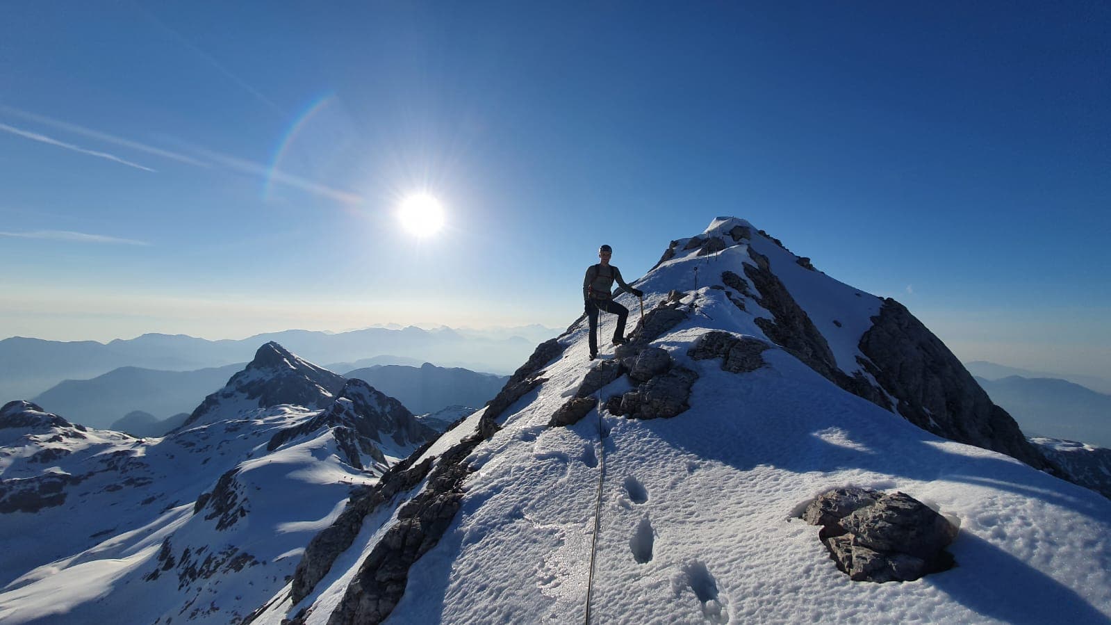 Winter climbing on Triglav ridge with mountaineer on snowy summit under bright sun.