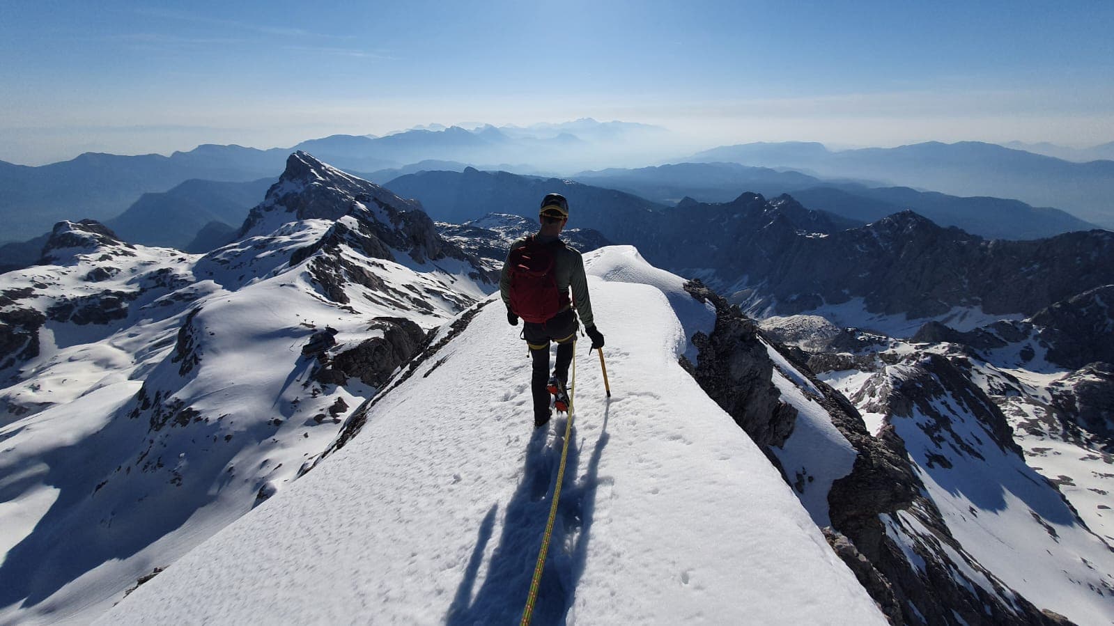 Mountaineer walking on snowy Triglav ridge with distant blue mountain ranges in winter.