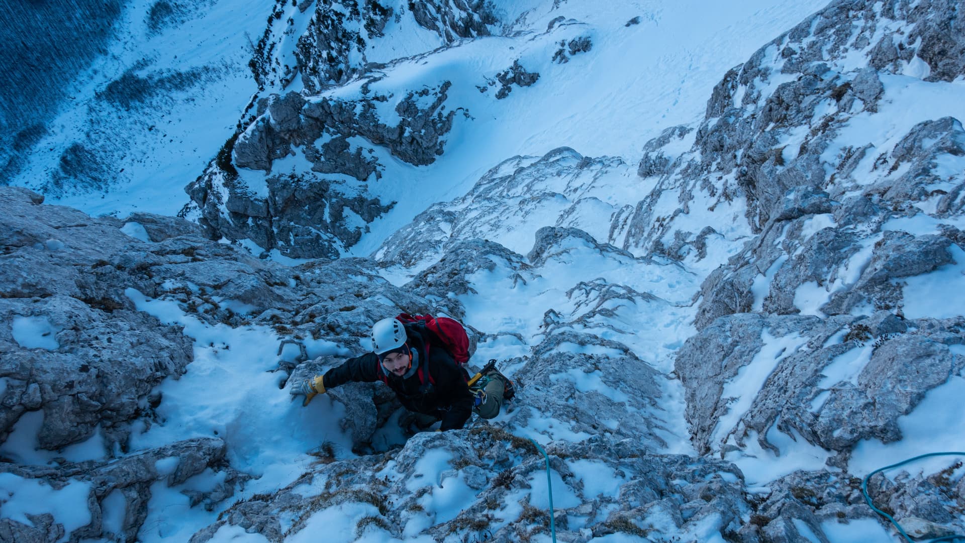 Mountaineer climbing icy, snow-covered rocks with a red backpack in winter.