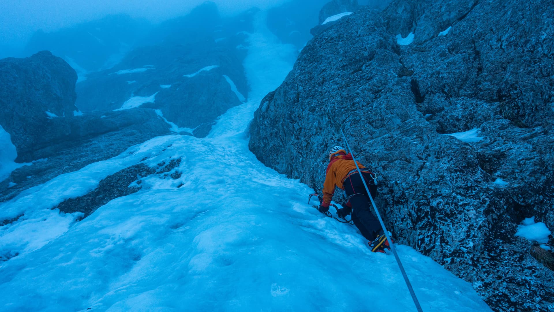 Ice climber ascending snowy, rocky alpine terrain in heavy fog or low light.