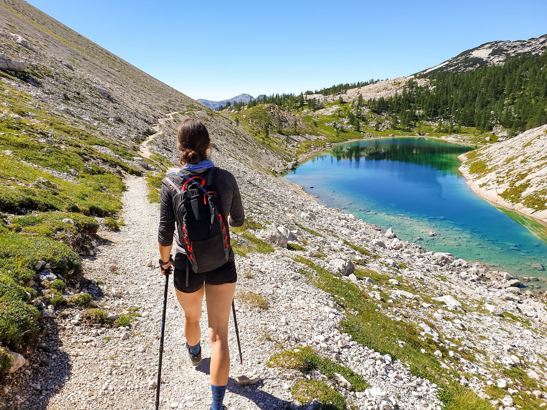 Hiker with backpack and poles walking on rocky trail above turquoise alpine lake in Julian Alps.