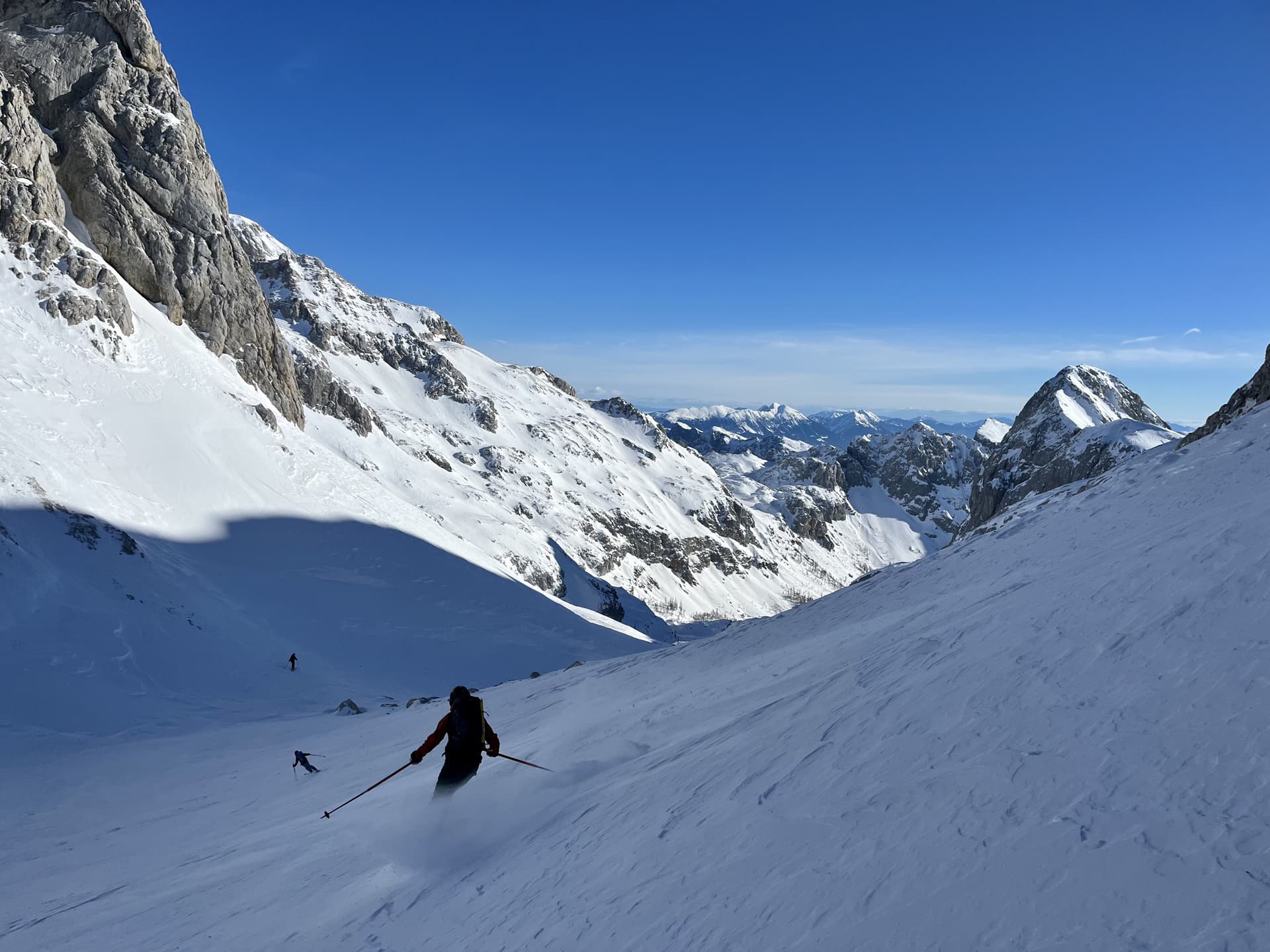 Ski touring down a steep, snow-covered alpine slope under a clear blue sky.