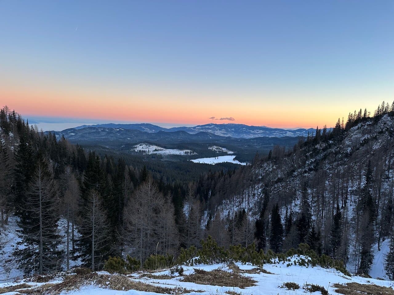 Snowy mountain forest landscape overlooking a valley and distant hills at sunset.