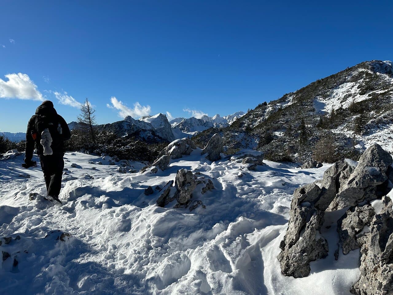 Hiker with backpack walking on snowy mountain trail toward snow-capped peaks under clear blue sky.