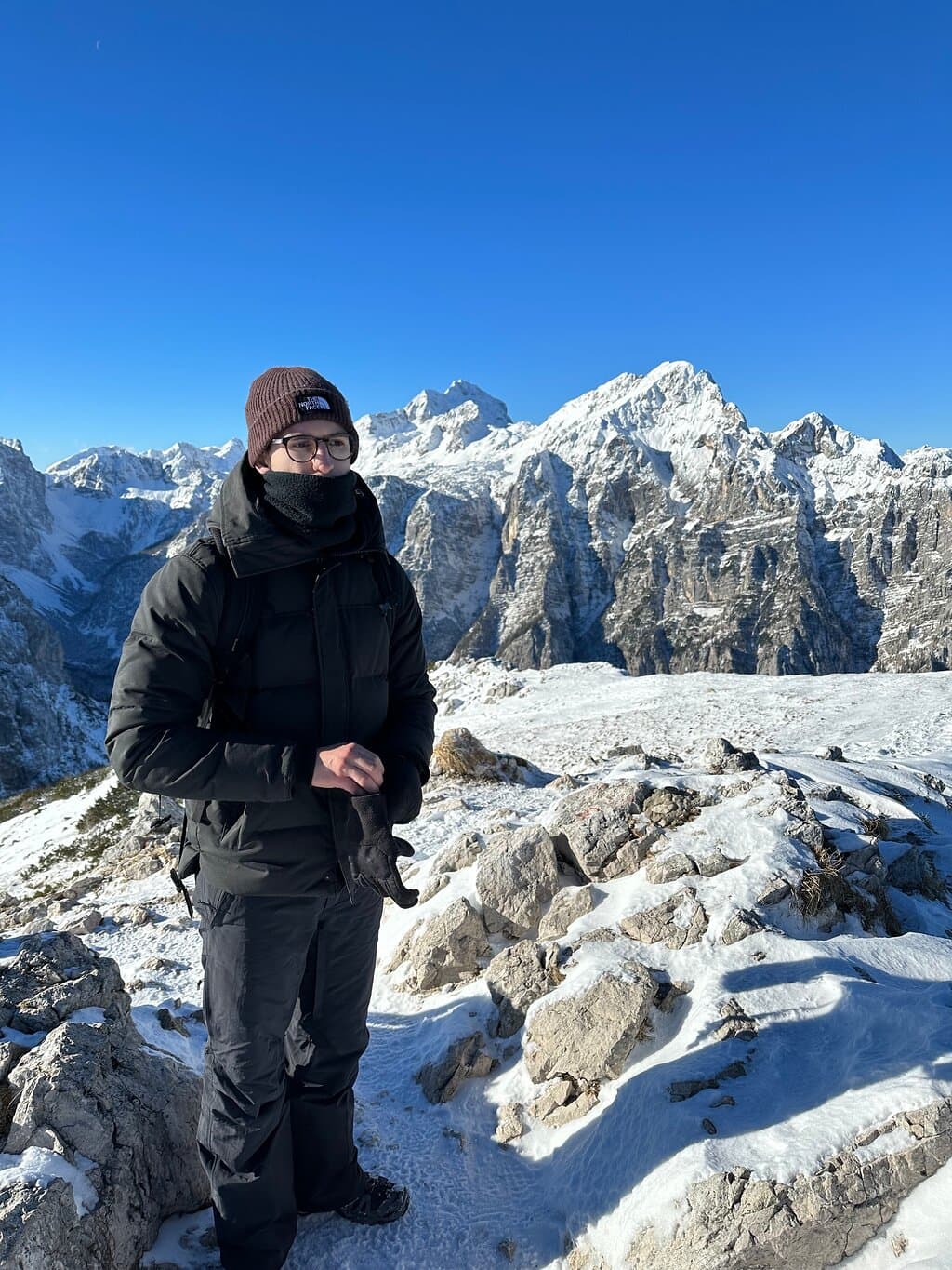 Hiker in winter gear standing on snowy mountain ridge with jagged, snow-capped peaks.