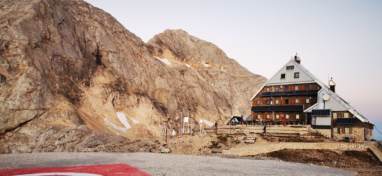 Mountain hut nestled against a large, rocky mountain face with patches of snow