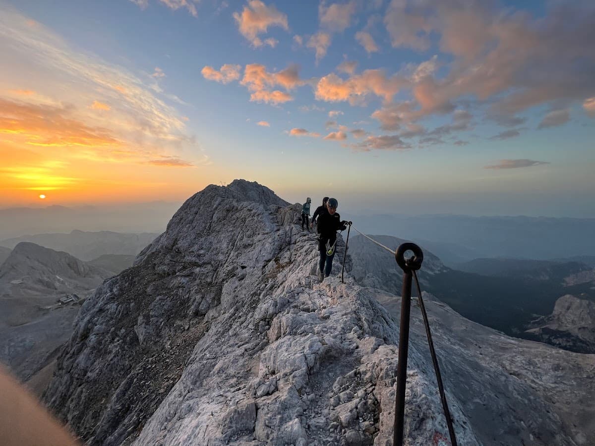 Hikers on rocky mountain ridge with safety cable at sunrise over distant peaks