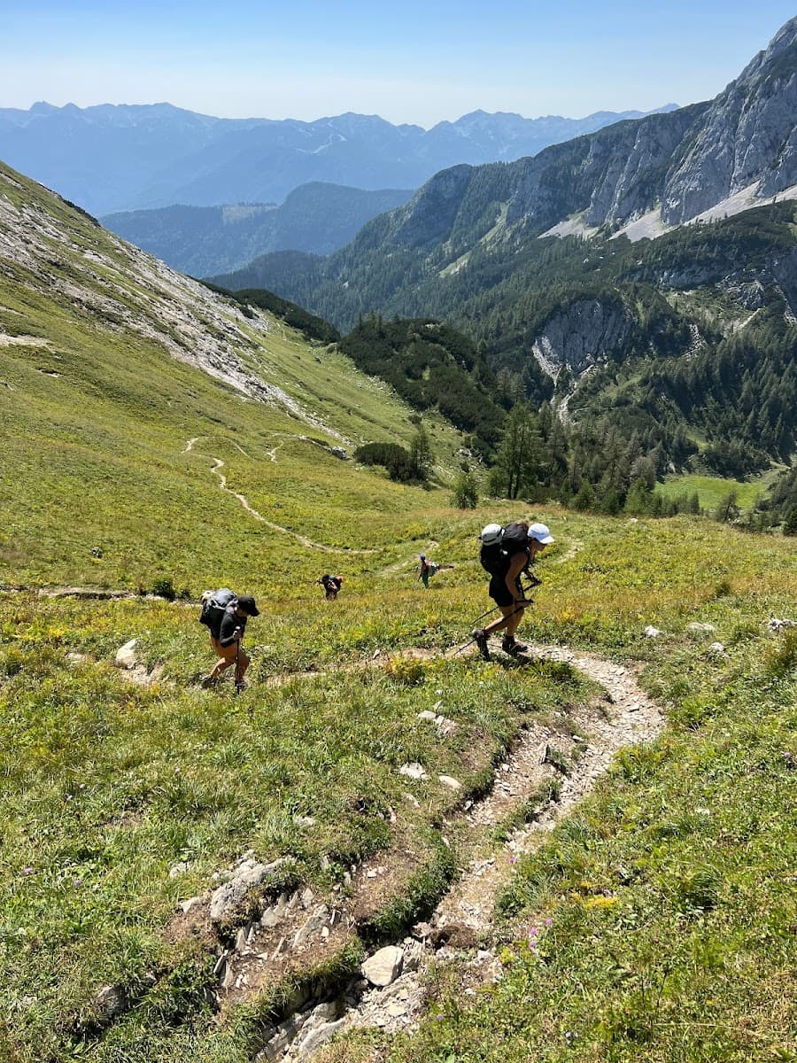 Hikers with backpacks ascend a grassy mountain trail with steep slopes and distant blue mountains.