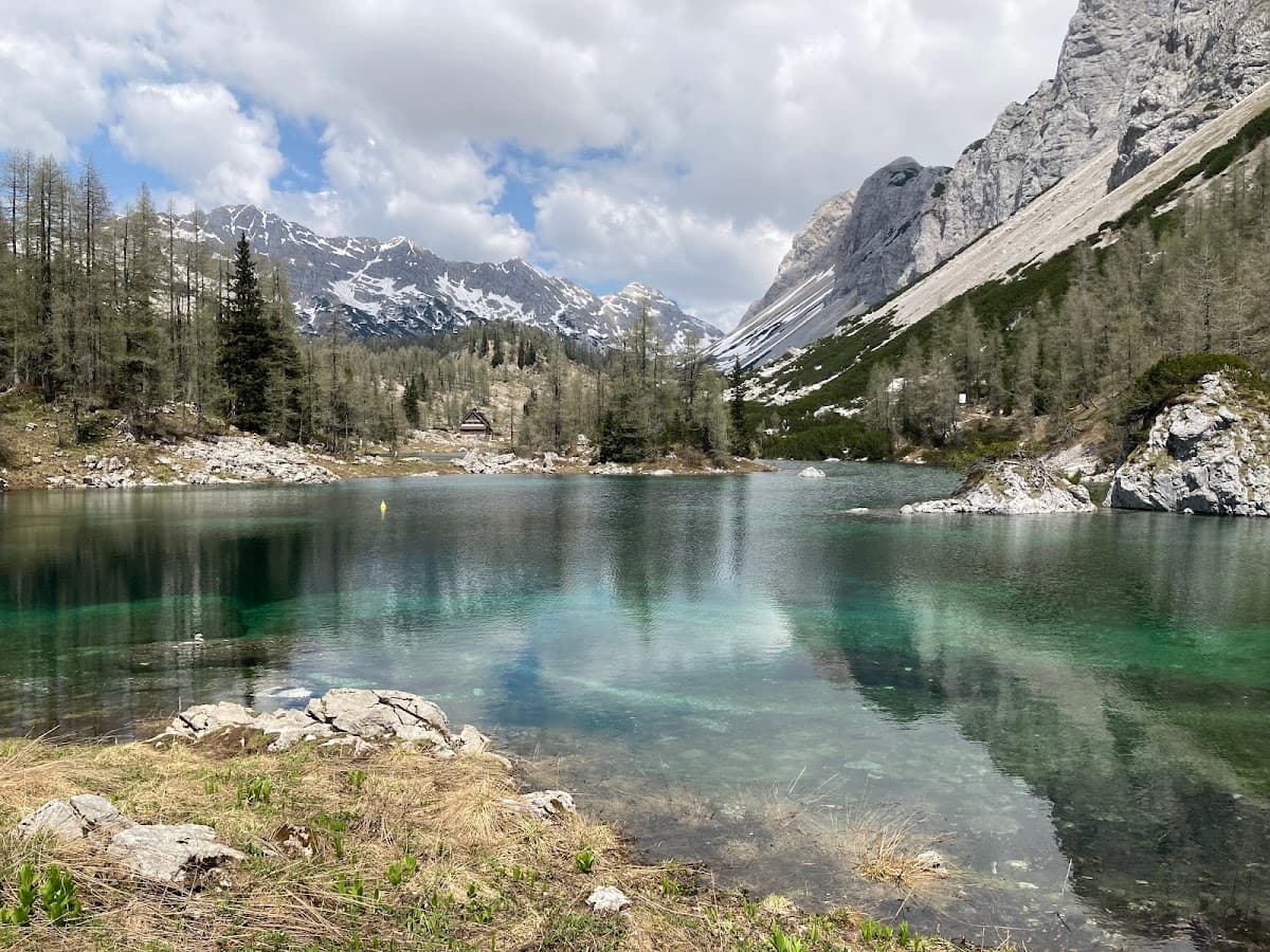 Alpine lake with clear turquoise water, snow-capped mountains, and pine forest under cloudy sky.