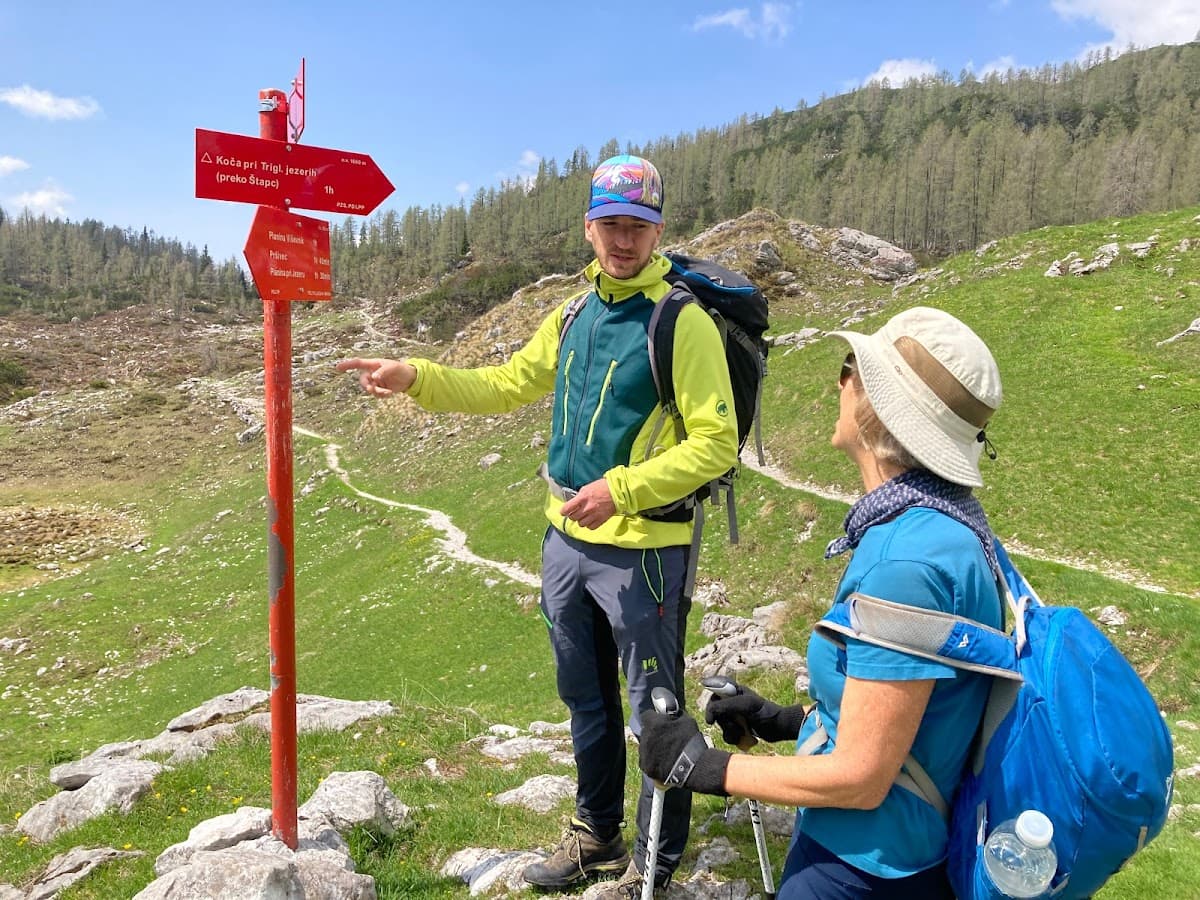 Hikers consulting a red trail marker on a grassy alpine slope with a winding path.