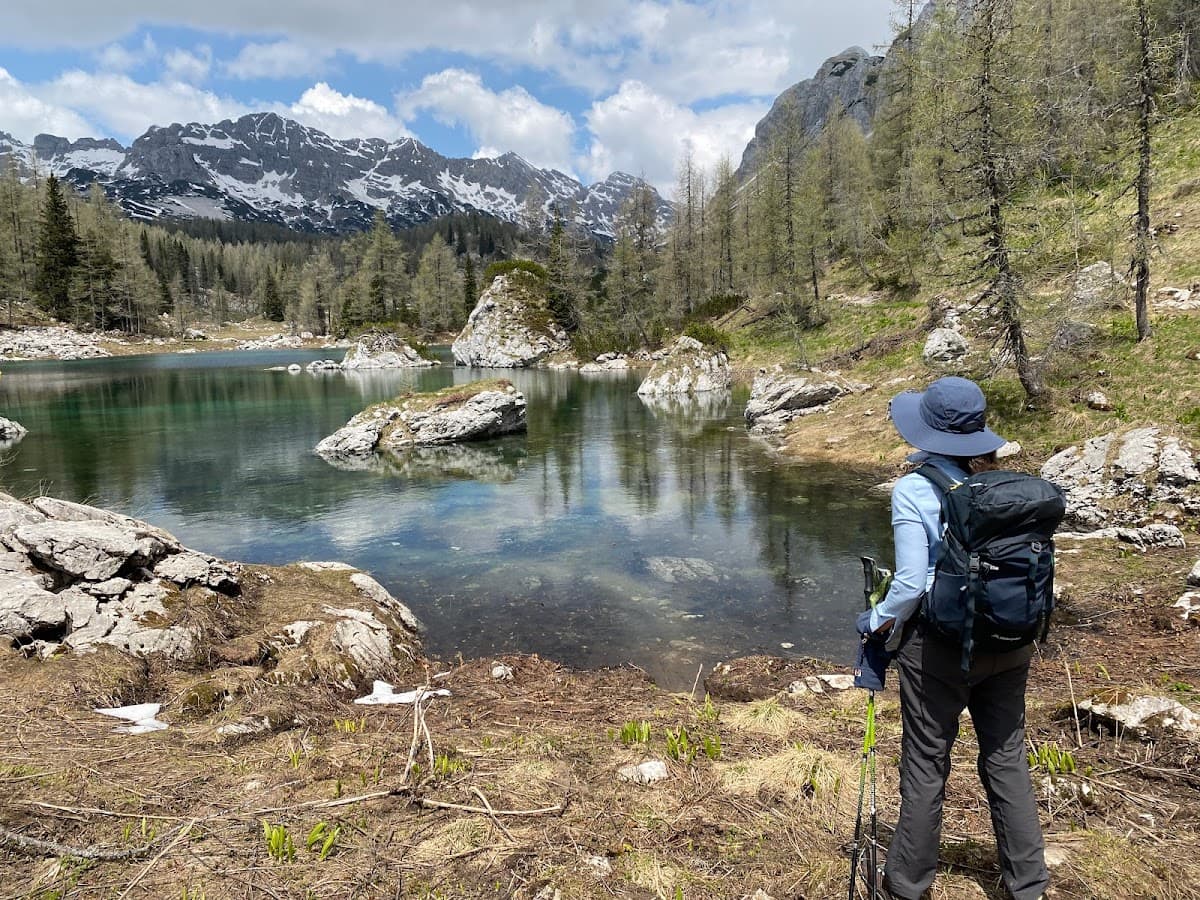 Hiker with backpack views clear alpine lake with snow-capped mountains and sparse spring trees.