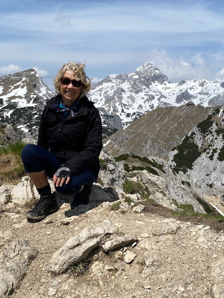 Hiker crouching on rocky mountain trail with snow-capped peaks in background