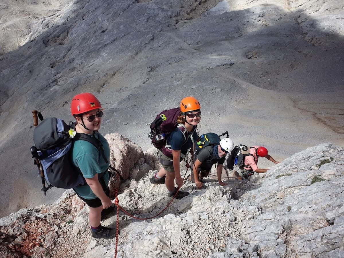 Hikers with helmets and backpacks ascending steep, rocky mountain terrain on a sunny day.