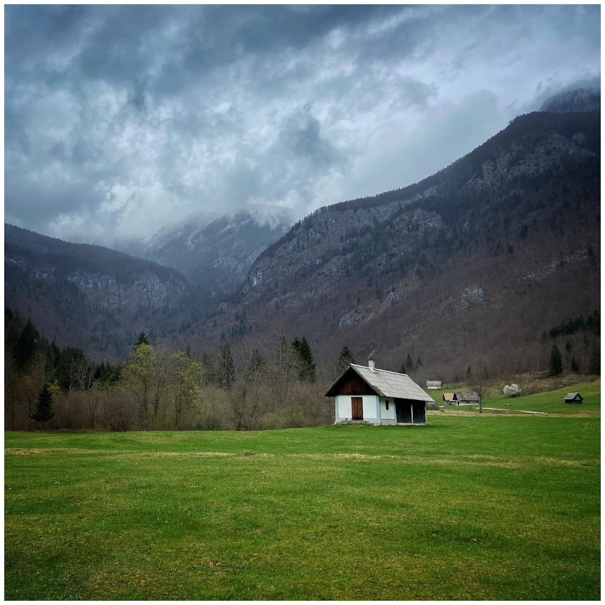 Small wooden cabin in a green meadow against steep, forested mountains under dark clouds.