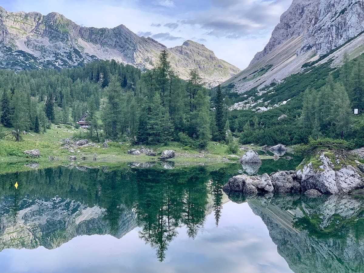 Mountain lake reflecting steep rocky slopes, pine forest, and a small cabin under cloudy sky.