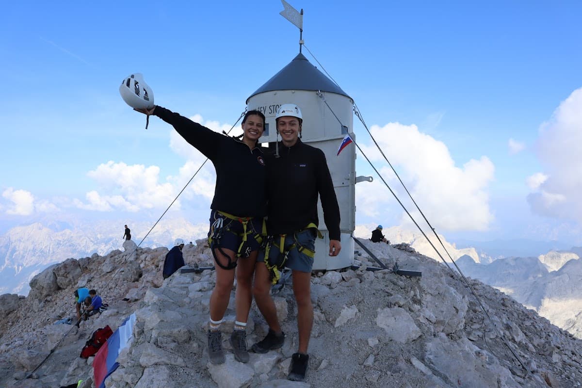 Two hikers with harnesses stand by a summit marker with a Slovenian flag on a rocky mountain peak.