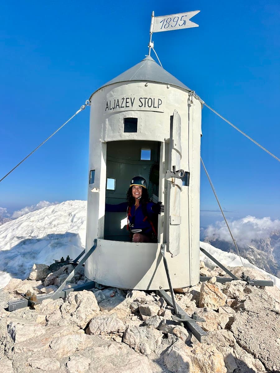 Hiker at Aljažev Stolp summit shelter on rocky peak with snow and blue sky
