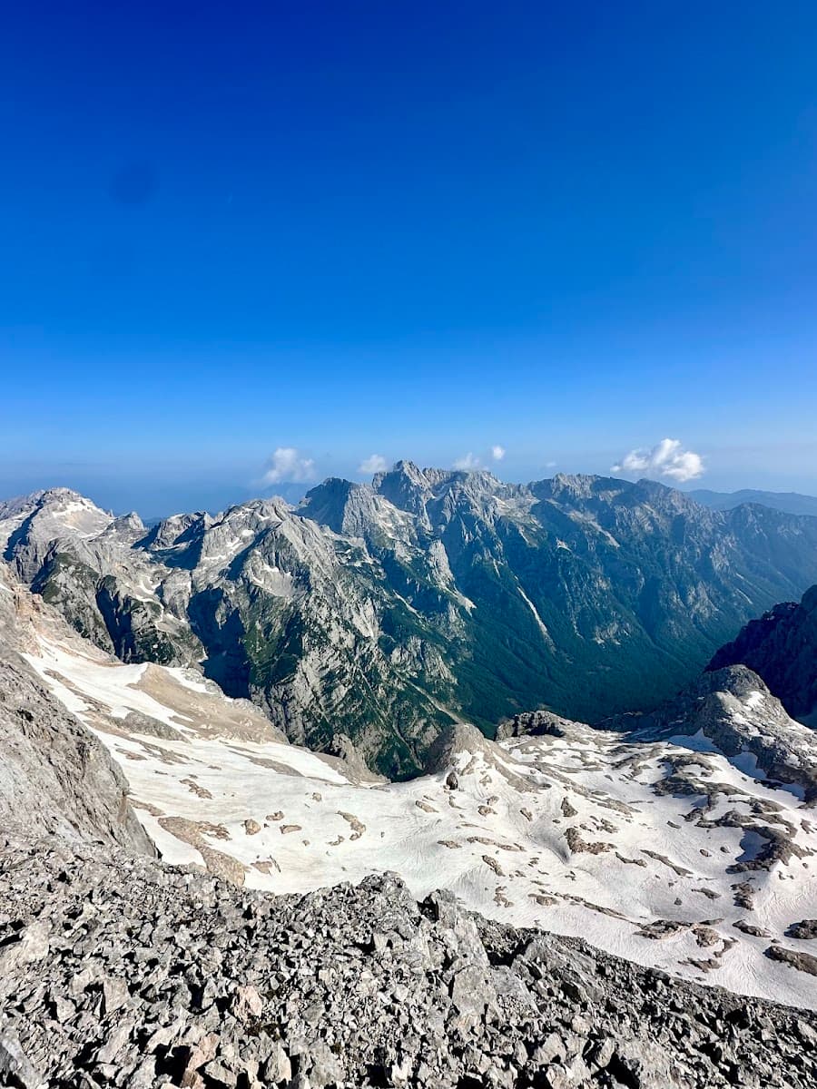 Rocky mountain summit with patches of snow overlooking deep valleys under a clear blue sky.