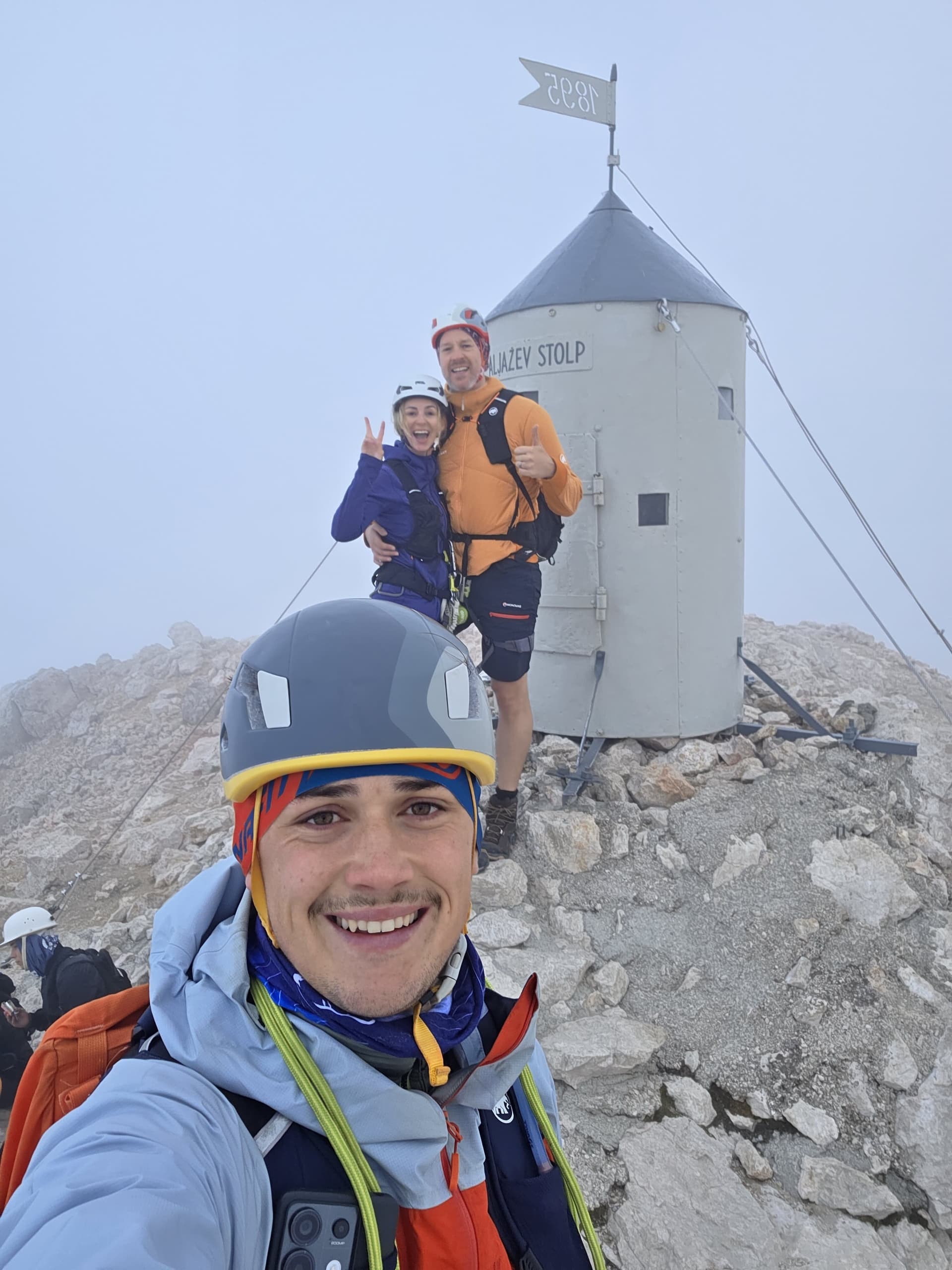 Climbers taking a summit selfie by Aljažev Stolp in foggy mountain terrain.