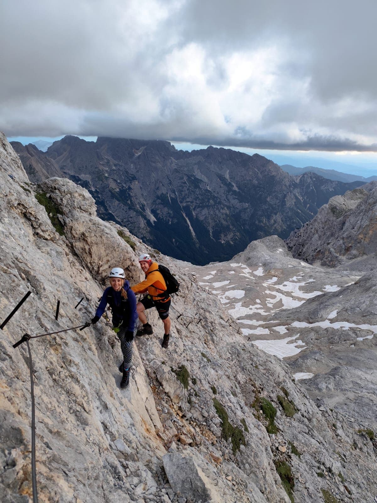 Hikers ascending via ferrata cables on steep rocky mountain slope with snow patches below
