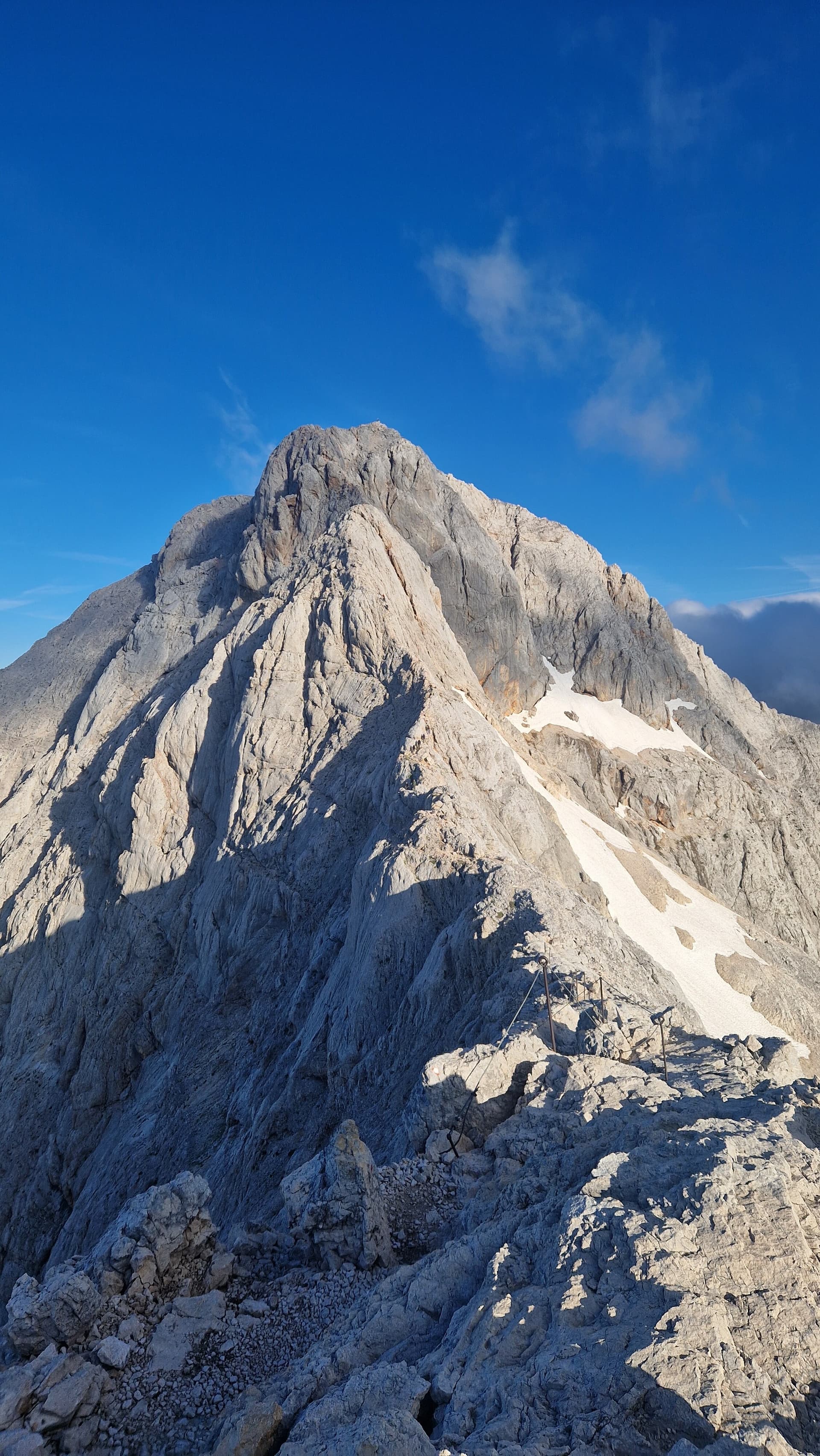 Rocky mountain peak with patches of snow under a clear blue sky, featuring climbing cables.