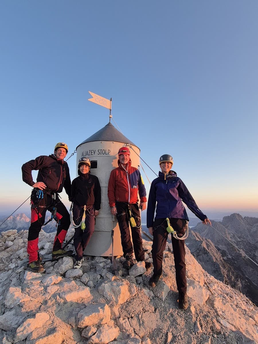 Four mountaineers with helmets stand by Aljažev Stolp on a rocky mountain summit at sunset.