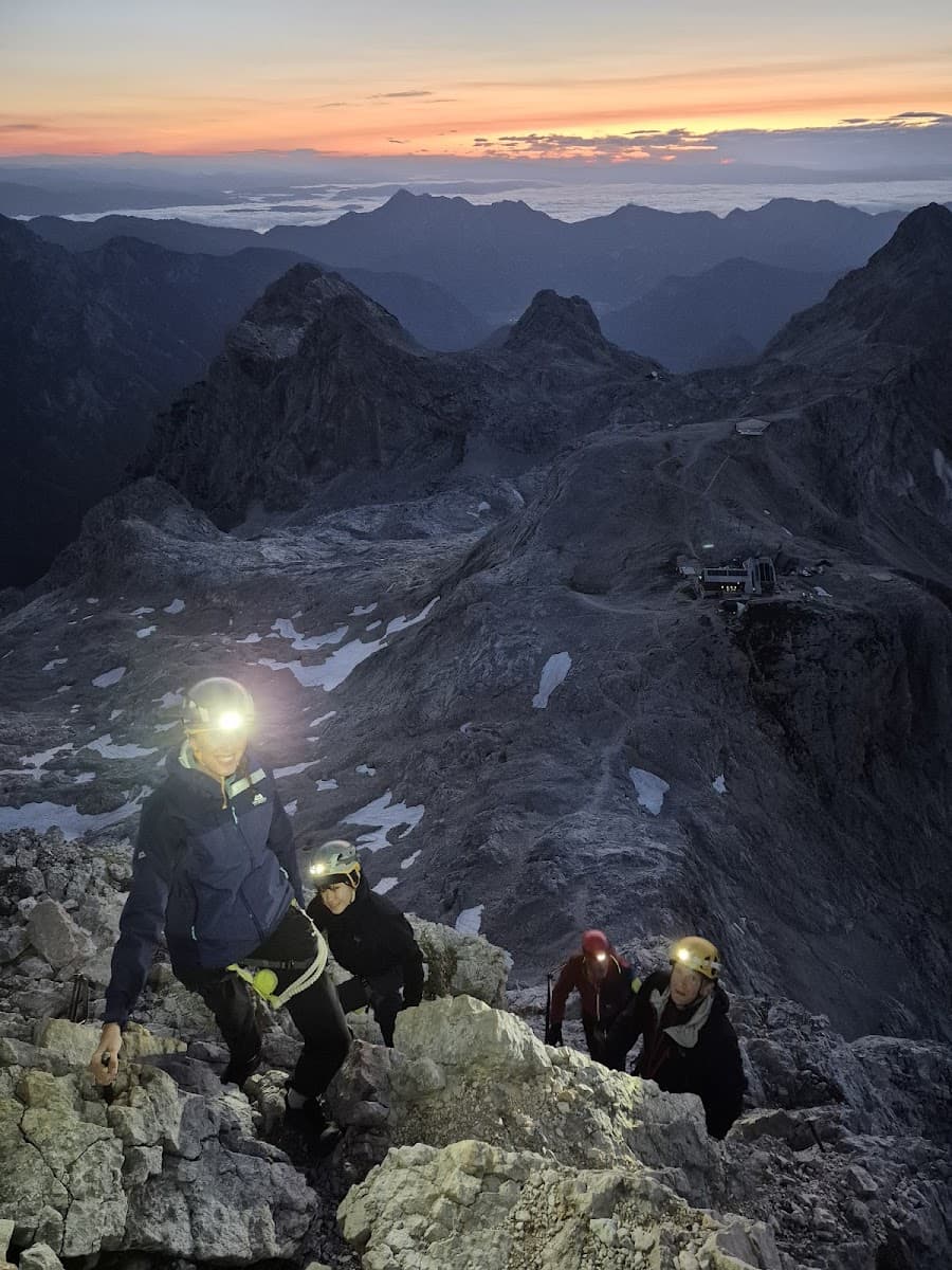 Hikers with headlamps ascending rocky terrain at sunrise over distant mountain ranges.