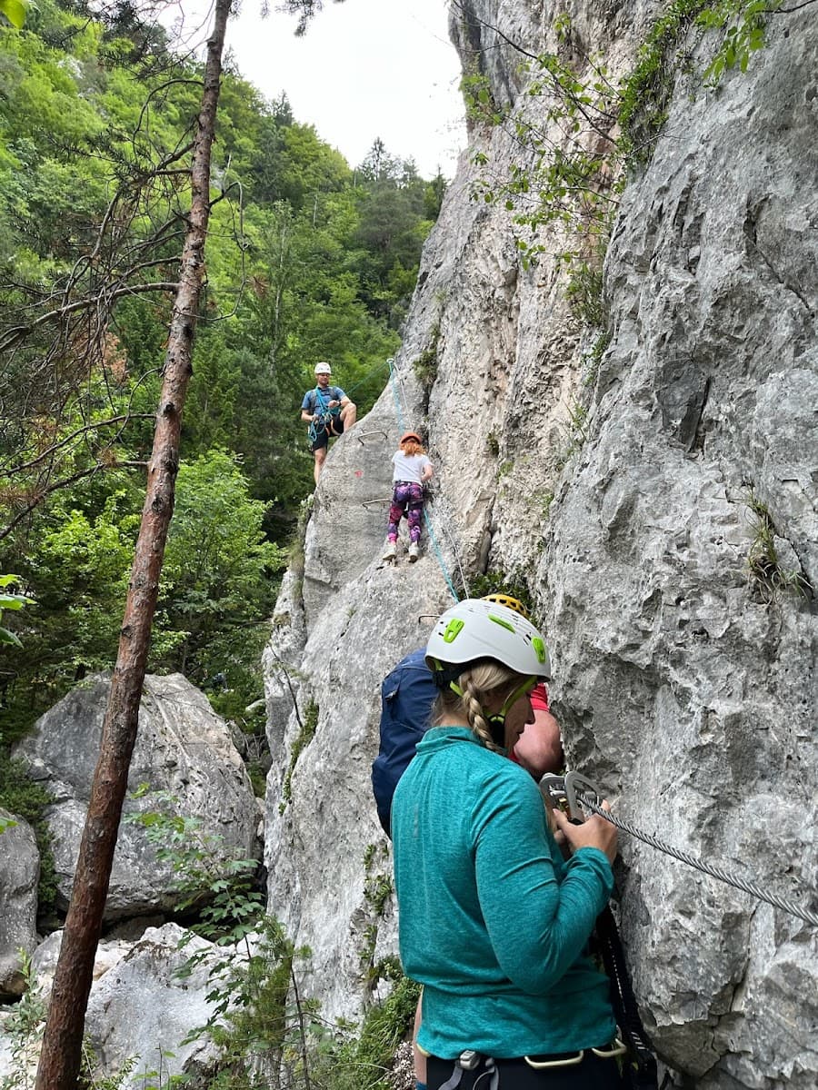 Climbers using via ferrata cables on a steep rock face surrounded by lush green forest.