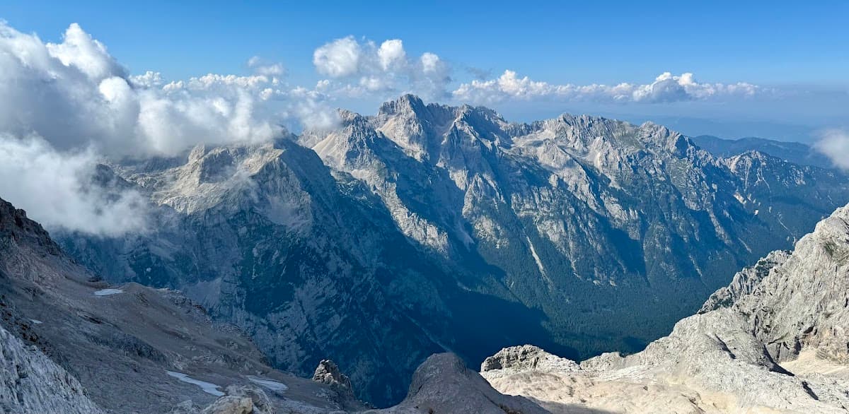 Rugged, rocky mountain peaks rising above dark green forests under a blue sky with white clouds.