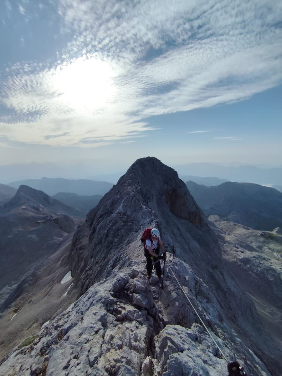Hiker with helmet ascending narrow rocky mountain ridge under bright sun and clouds