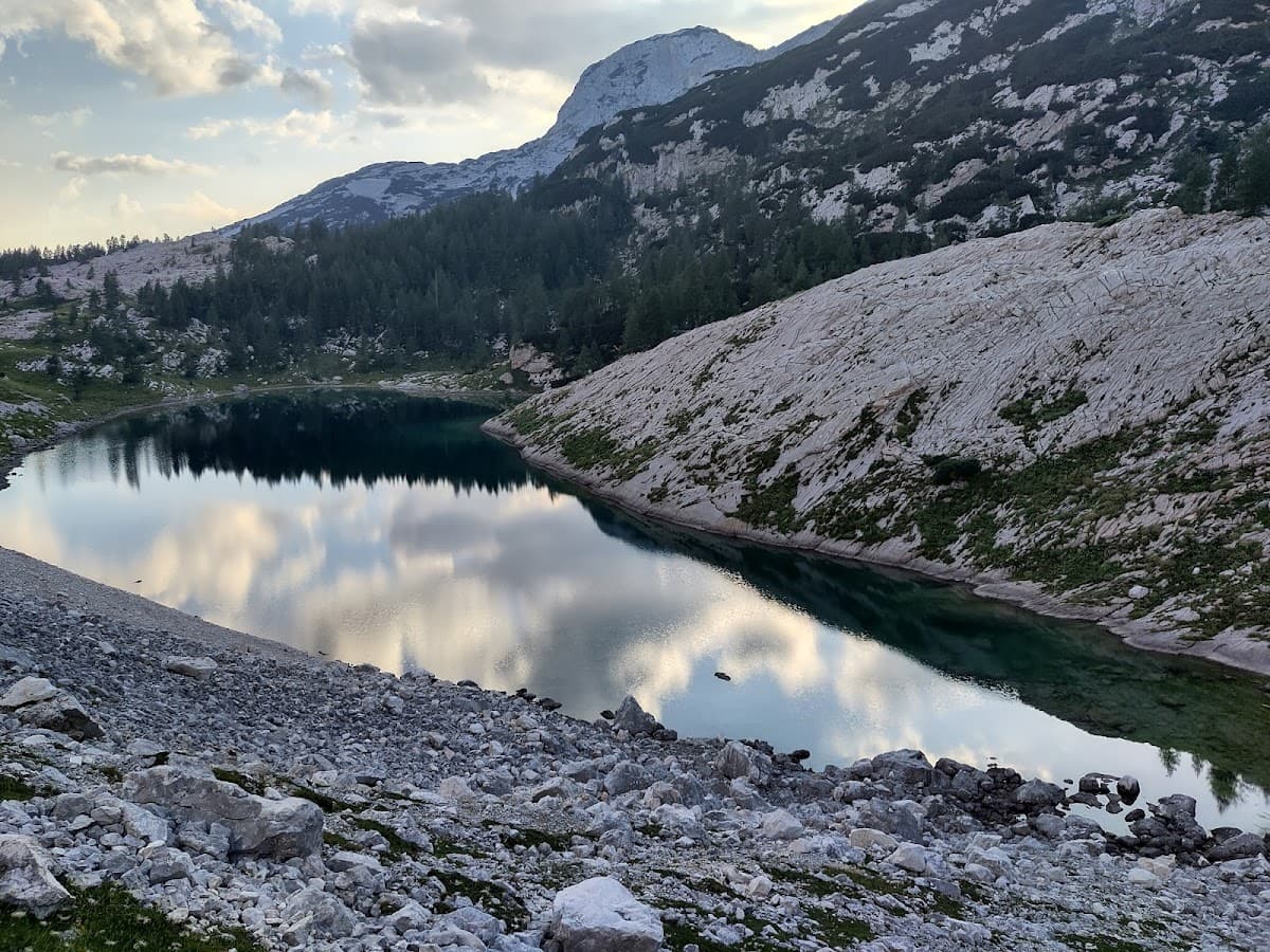 Alpine lake reflecting clouds and surrounding mountains with rocky shore and scattered pine trees.