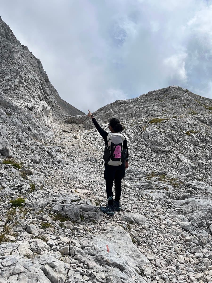 Hiker pointing upward on rocky mountain trail under cloudy sky