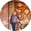 Man standing indoors near brick wall and large, ornate burgundy and gold patterned rug.