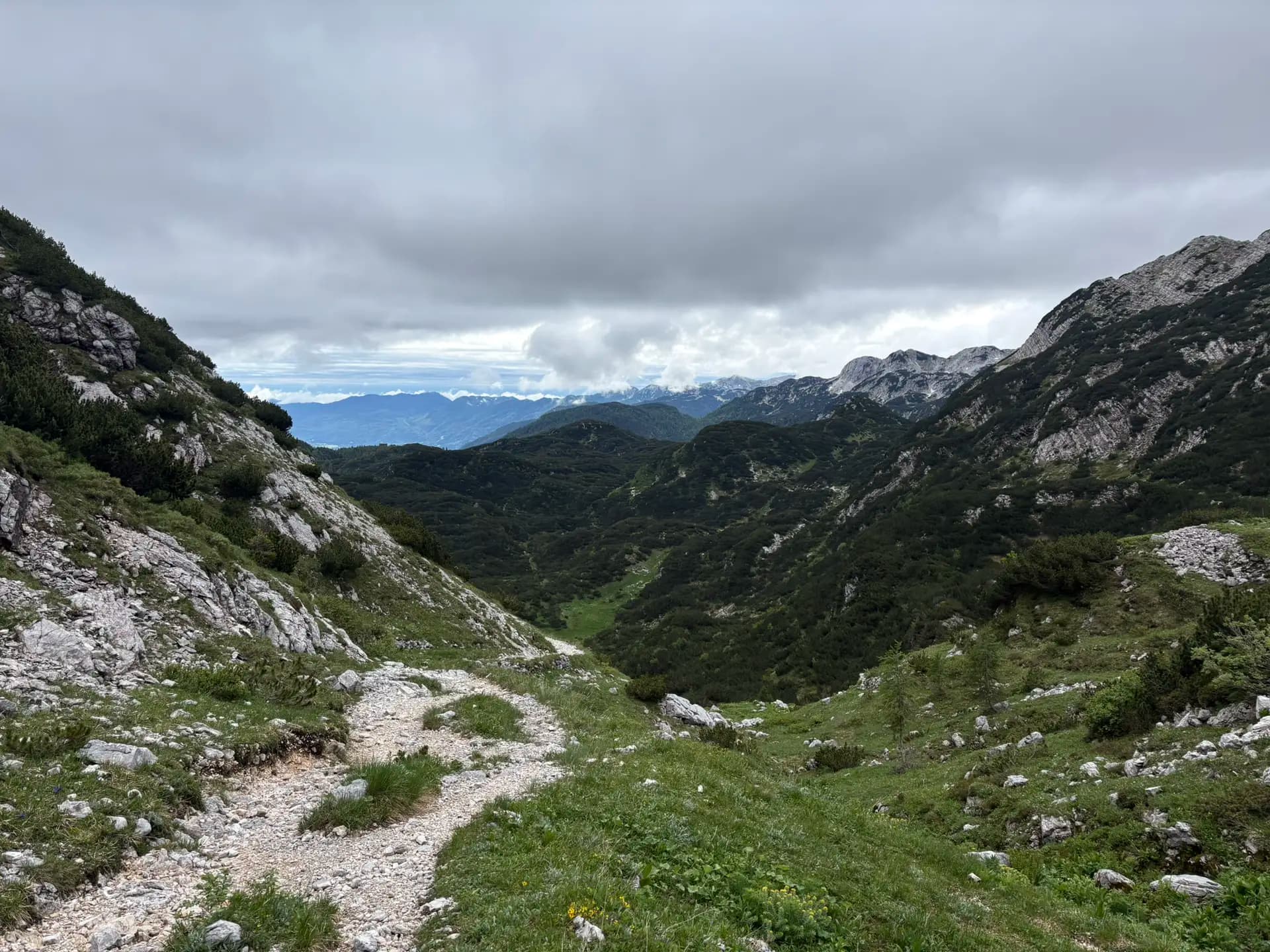 Rocky hiking trail winding through the Julian Alps with green mountain slopes and a cloudy sky.