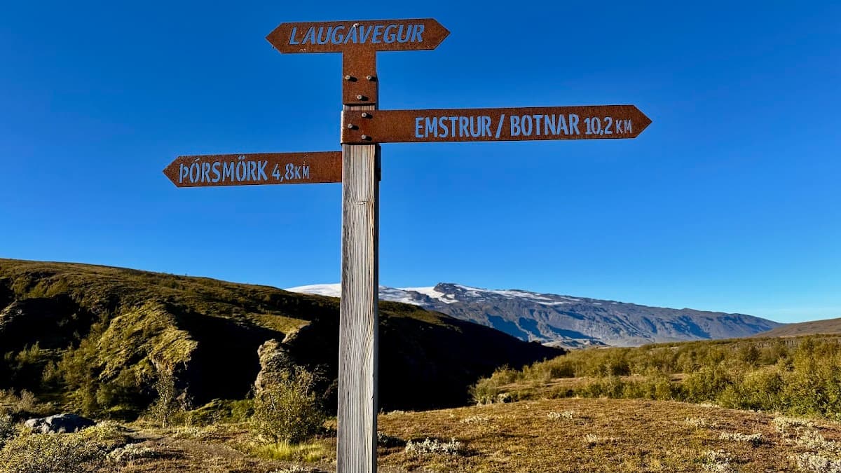 Hiking trail sign with directions to Laugavegur, Þórsmörk, and Emstrur against a mountainous landscape and blue sky