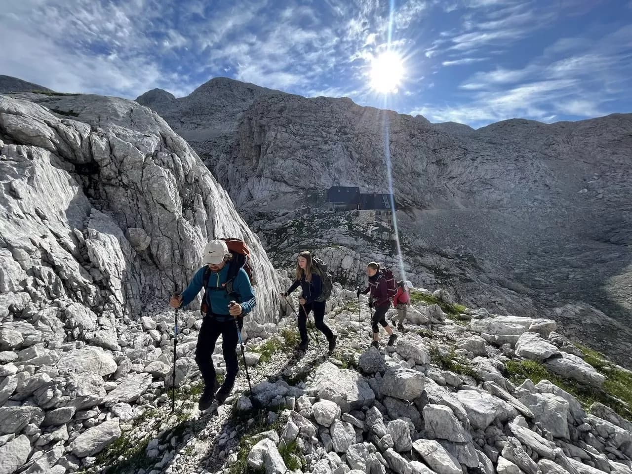 Hikers ascending rocky terrain toward a mountain hut under bright sun and blue sky