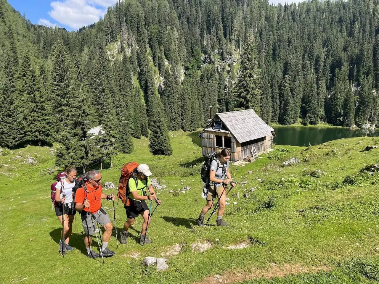 Hikers with backpacks and poles trekking near a wooden cabin by a mountain lake.