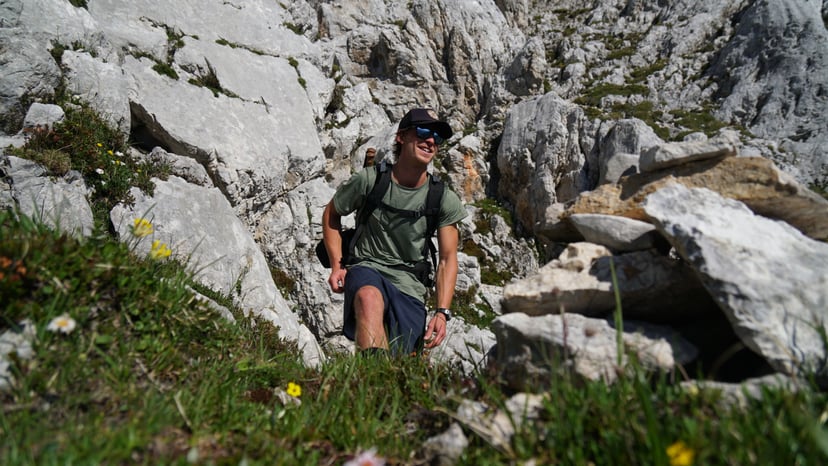 Hiker with a backpack walking on a rocky mountain trail with alpine flowers.