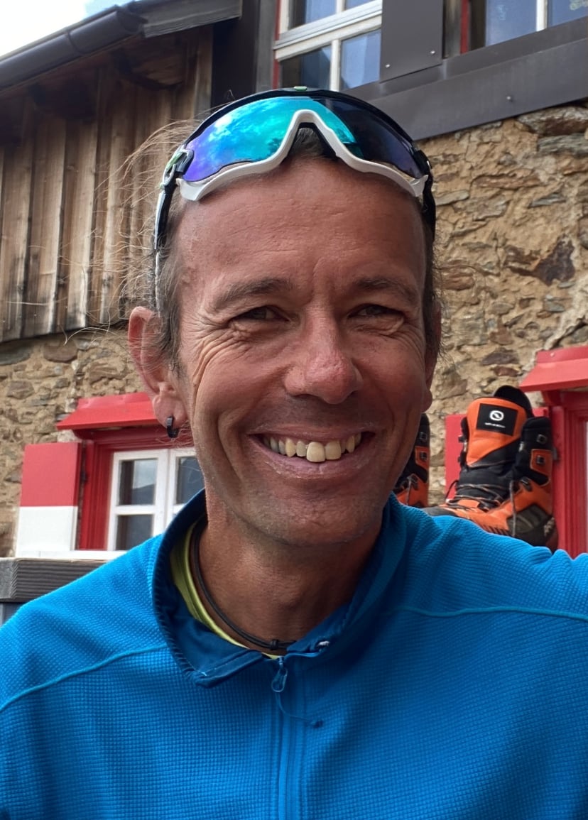 Smiling man wearing a blue fleece and sports sunglasses in front of a rustic stone building.