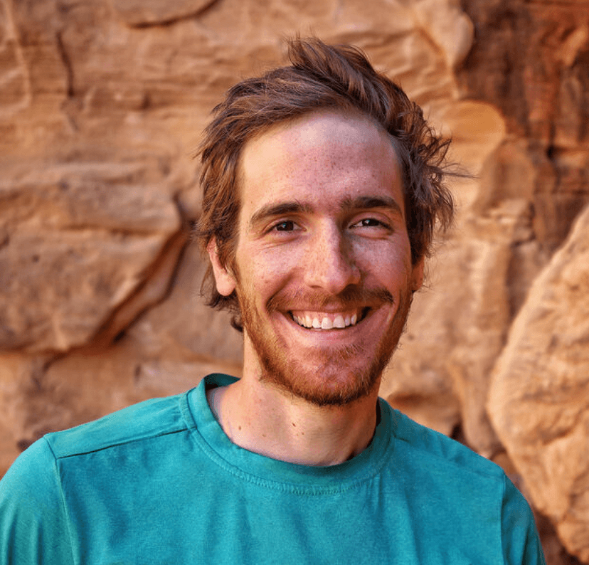 Smiling man with a beard wearing a teal shirt in front of a rock wall.