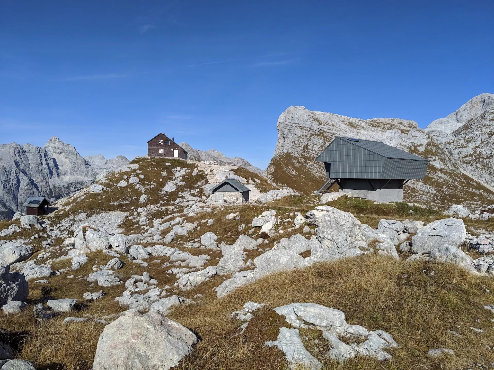 Prehodavci lodge and bivouac huts on rocky, grassy alpine terrain under a clear blue sky.