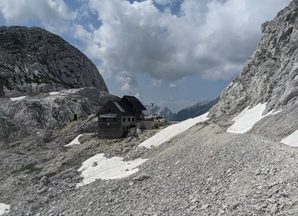 Mountain lodge nestled in rocky terrain with patches of snow under a cloudy sky, Dolič Mountain.
