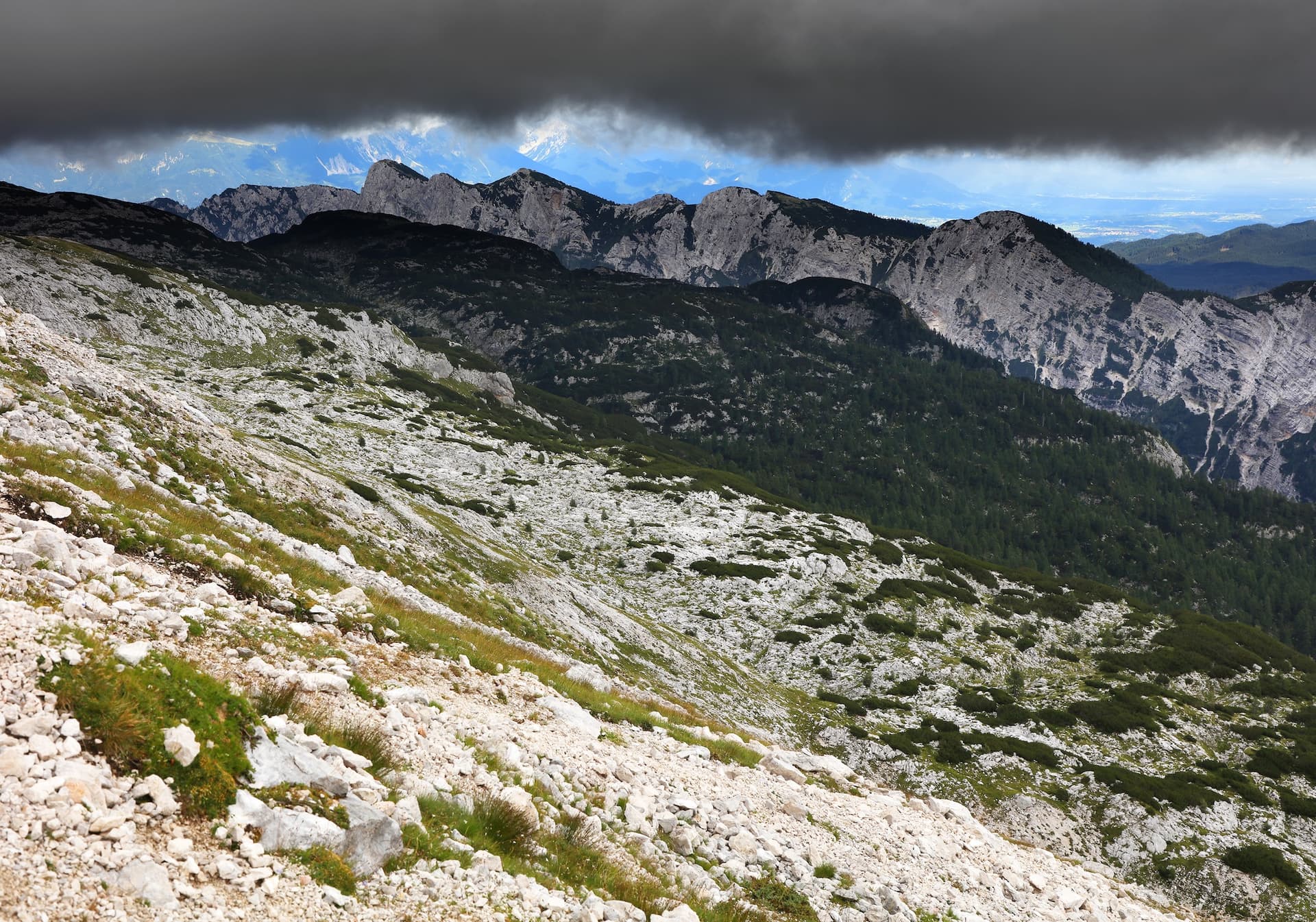 Rocky alpine slope with sparse grass leading to dark mountains under stormy clouds