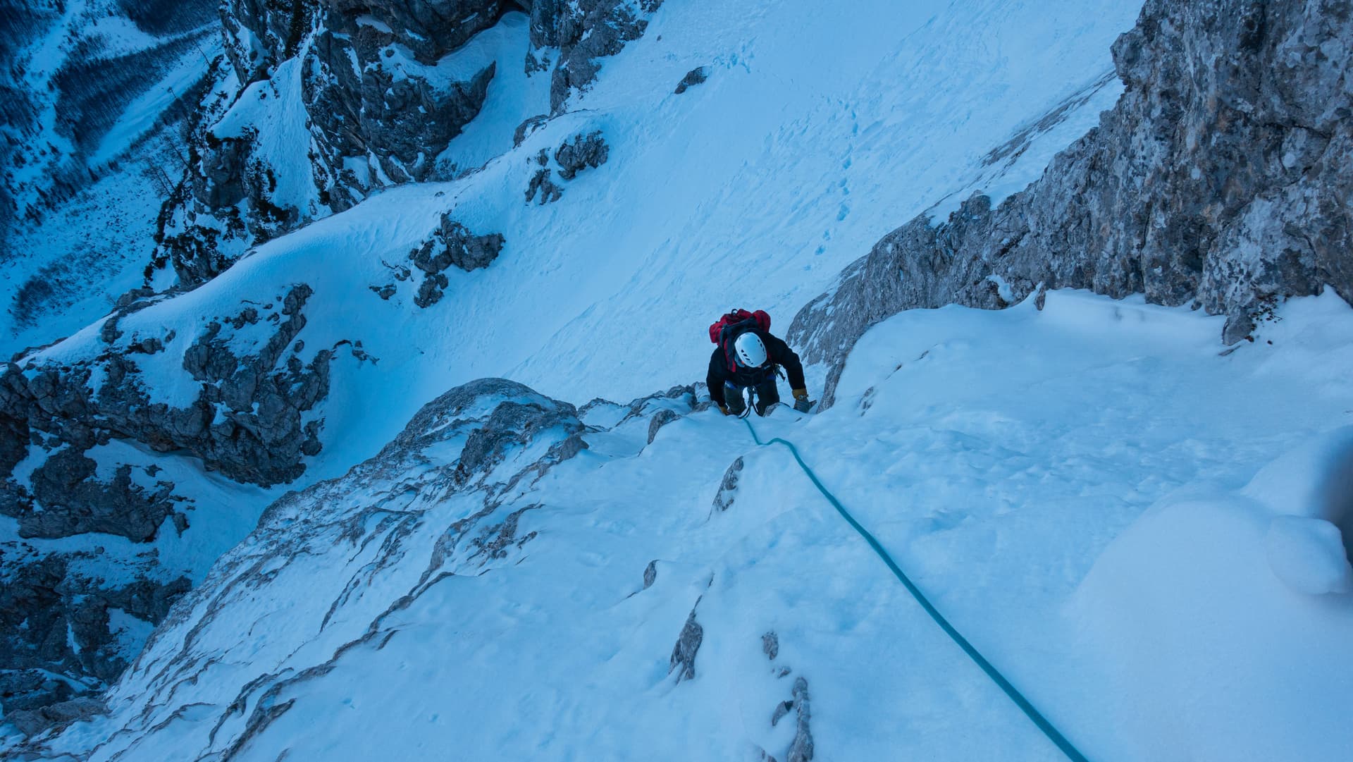 Mountaineer climbing steep snow and rock face with ice axe and rope in blue twilight.