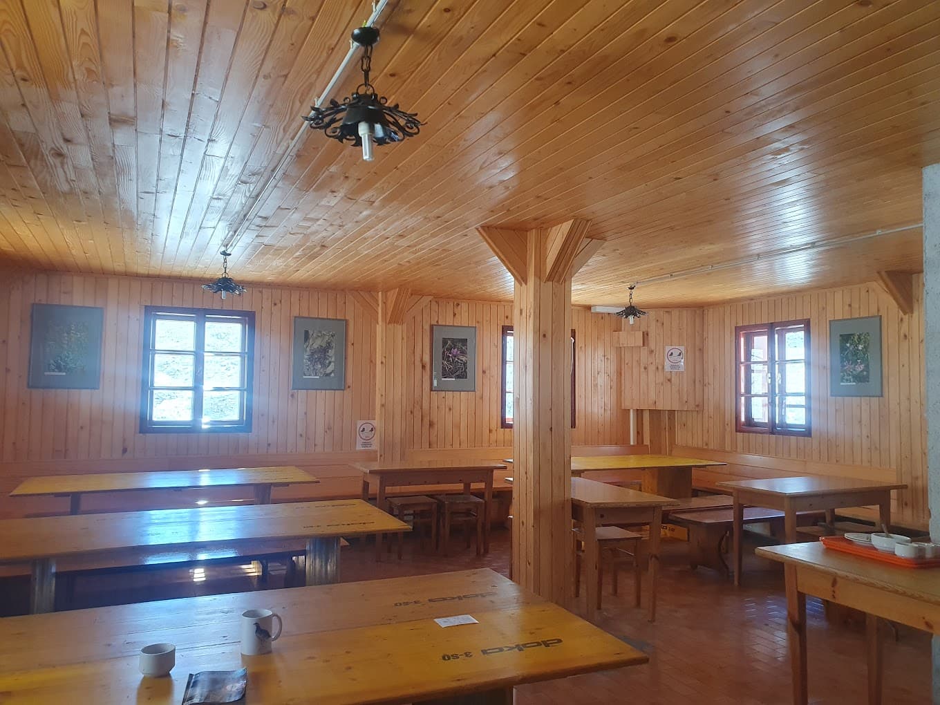 Wooden interior of a mountain hut dining hall with long tables and windows showing bright daylight.