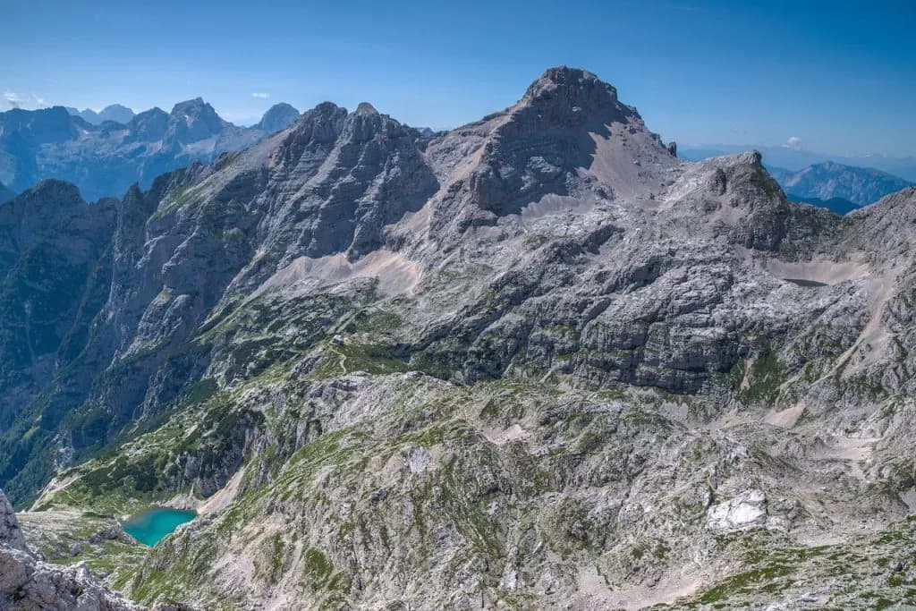 Rocky mountains with sparse vegetation and a small turquoise alpine lake under a clear blue sky.