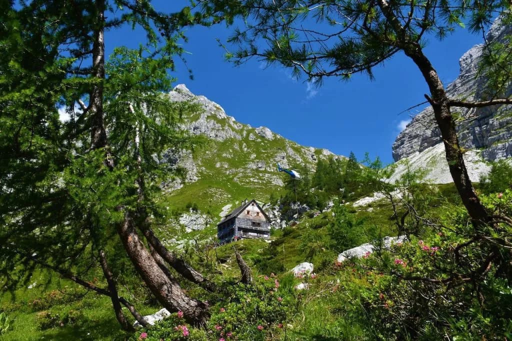 Helicopter supplying Vodnik lodge nestled in green mountainside under blue sky