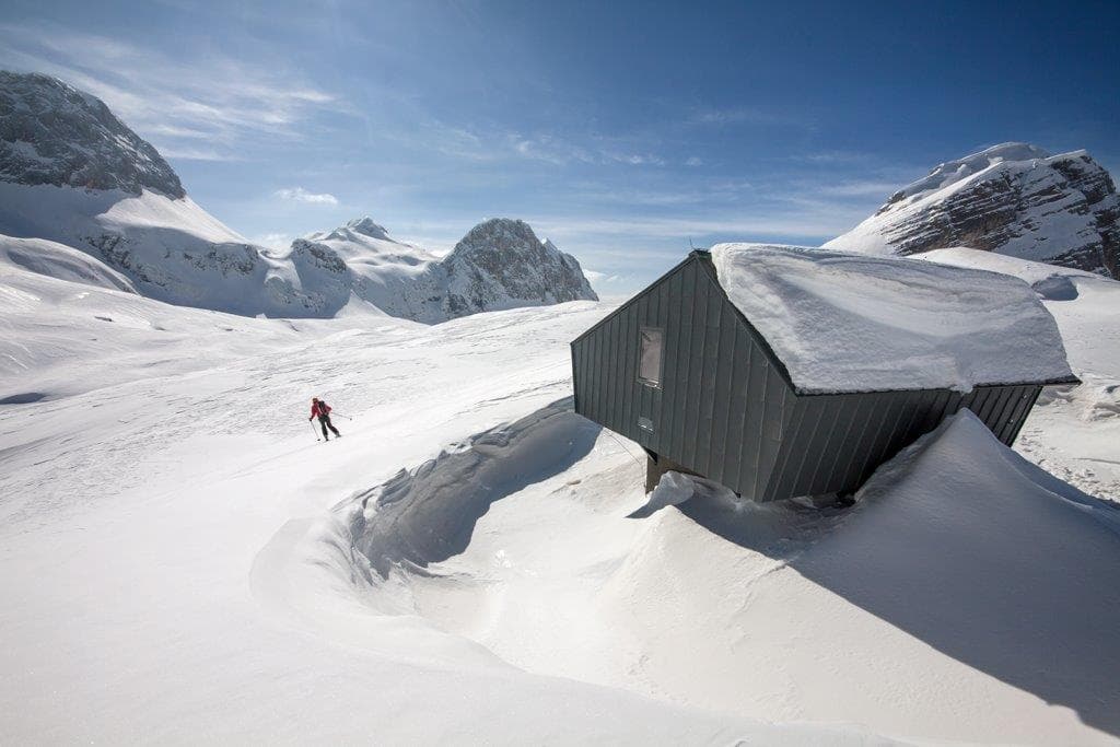 Skier descending snowy slope near modern mountain bivouac hut under blue sky.